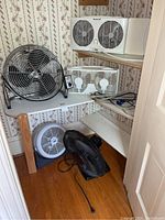 Photo of large Duracraft fan with metal grill on a shelf along with smaller white box fan, black and white small personal fans on floor.