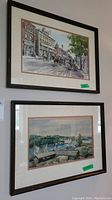 Two framed prints hung on a wall, upper one showing horse-drawn carriages and town buildings, lower one showing sailboats on a river.