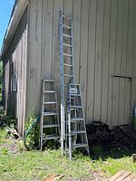 Full view of three ladders leaning against outer wall: one tall extension ladder and two smaller step ladders.