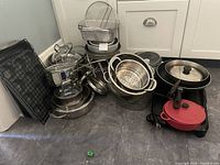 Overview of assortment of pots, pans, baking trays and colanders stacked together on floor near cabinets.