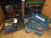Mastercraft router box, Makita sanders in cases, drill bits, saw blades, belt sander, Skilsaw and additional hand sanders displayed in a shelf.