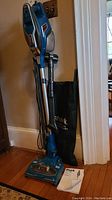 Full view of the blue Shark Rocket Duo Clean upright vacuum with power cord and accessory bag, standing next to a wall and door.