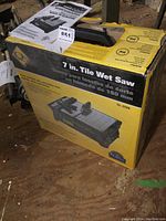 Photo of the boxed 7 inch tile wet saw with manual on top, showing front and side of box with product image and branding