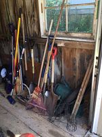 Full view of the assorted yard tools including shovels, rakes, broom, car brush, and watering cans leaning against wooden shed wall.