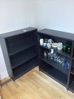 Pair of black bookshelves positioned in corner of room; one bookshelf contains various glassware and mugs; visible adjustable shelves with peg holes.