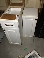 Two white kitchen cabinets side by side on basement floor, showing fronts and tops with unfinished wood edges.