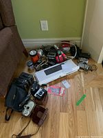 Overview photo showing cameras, keyboard, phone, flashlight, cassette player, cords on wood floor near wall.