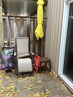 Photo showing the outdoor zero gravity chair with beige fabric, yellow folded umbrella and brown base, two small resin side tables (brown and red), and two bags of mulch on the ground near a patio door and wood fence.