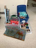 Three large plastic totes with assorted Christmas decorations, wrapping paper, and gift bags visible, along with a blue decorative mat with moose and bird illustration.