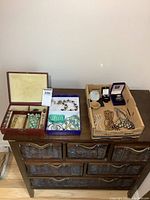 Photo shows a wooden jewelry box containing various bracelets and necklaces including turquoise beads, alongside two cardboard boxes filled with assorted costume jewelry such as necklaces and boxed rings or brooches.