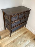 Angle view of wood dresser with wicker drawers and unattached wood top, placed against a wall on light wood floor.