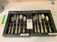 Two unopened boxes of Paderno flatware sets placed side by side, each box displaying 5 utensils including forks and spoons with polished stainless steel finish.