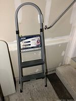 Full view of the Cosco folding step stool standing upright, showing the two gray plastic steps, metal frame, and rounded handle with label.