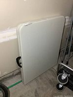 Folded resin folding table resting against a wall, showing the handle and thickness.