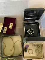 Clear plastic container with small jewelry boxes containing enamel cuff links, snake chain bracelet, silver-tone cuff links, tie bars, purple stone and gold band ring