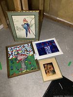 Overview of four framed artworks on floor against wall showing all pieces in the lot including parrots painting, green background bird painting, abstract floral oil painting, and sunflower print.