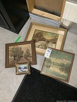 Four framed artworks laid on ground including two oil on board paintings of winter scenes and two older landscape prints.