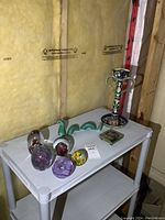 White shelf holding five colorful glass paperweights, green ceramic Loch Ness monster figurine, and tall candle holder with marble base