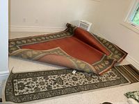 Large rust-colored rug with decorative leaf border partially rolled, with runner and small rug nearby on carpeted floor
