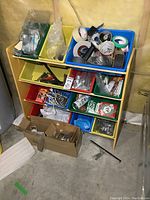 Front view of wooden shelf with colored plastic bins containing rolls of tape, tools, hardware items, wires, and a cardboard box below.
