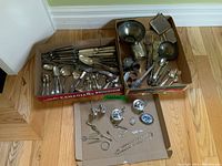 Photo showing the assortment of silver plate cutlery laid out in two cardboard boxes and flat cardboard with small silver items