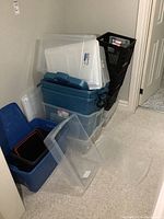 Stack of various sized plastic storage totes and milk crates leaning against a wall, mostly blue and clear with a few black crates.