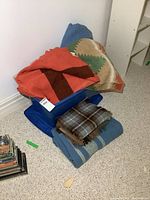 Four wool blankets and throws stacked on blue container in carpeted room corner; orange/brown blanket on top, green/beige one partially visible, blue blanket underneath, and plaid throw at bottom.