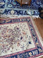 Partial view of both rugs overlapped on wooden floor, showing blue floral rug underneath and beige tree-of-life rug on top with fringe visible