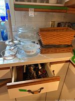 Kitchen counter with glass casseroles, Pyrex bowls, 9x13 dish, woven baskets, and kitchen drawer slightly open showing kitchen tools.