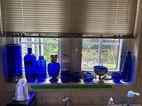 Wide view of various blue glass items arranged on a kitchen windowsill including candleholders, jars, bottles, and the silverplate dish with blue insert, showing overall lot