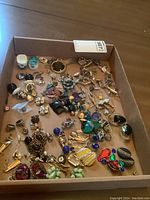 Top-down view showing a wide selection of jewelry including earrings, cufflinks, and pins in a box, displaying color and design variety.