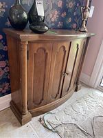 Solid wood hall cabinet with curved front, classical column side supports, two panel doors with round knobs, and top surface with visible wear and scratches.