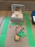 Photo showing two necklaces: a gold filled heart-shaped locket on a chain and a silver chain with pearl pendant, as well as a small ring box.