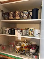 Full kitchen cupboard shelf showing four decorative mugs with painted outdoor scenes, various other mugs, blue ceramic pitcher, Pyrex liquid measuring cups, ceramic teapot set with warmer, and metal measuring cups and spoons.