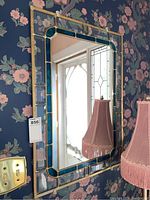 Overall view of the rectangular mirror hung on floral wallpaper showing the stained glass border and reflection of nearby window and lamp.