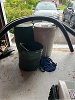 View of green and beige rain barrels with blue garden hose and green yard cleanup bag in front.