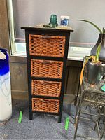 Front view of black wood stand with four wicker drawers beside a tall blue and white glass vase.