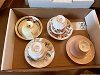 Four assorted teacups and matching saucers in a cardboard box showing different floral designs and colors