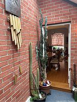 Full height view of the artificial tree cactus standing about 100 inches tall against a red brick wall, showing the plastic pot at the base and other potted plants nearby.