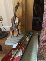 Violin displayed on brass-look stand with red-lined hard shell case open showing bow and rosin bag inside.