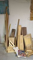 Wood boards and strips stacked against a wall, some leaning upright, showing natural wood, various sizes and thicknesses