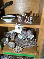 Full view of mixed floral chintz ware china items including cups, saucers, small dishes, a creamer jug, and spoon rests arranged on wood shelf.