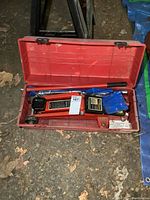 Red hydraulic trolley jack in a closed red plastic carrying case with black latches, lying on the ground.