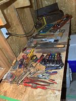 Wide view of various hand tools laid out on a workbench showing many pliers, wrenches, screwdrivers, files and hammers, and toolbox in background.