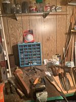 Photo showing an assortment of hammers, crowbars, files, nail sets, and a compartment caddy filled with nuts and bolts on a wooden surface under a shelf with jars containing nails and hardware.