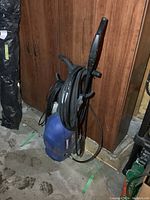 Side view of blue Simoniz 1500 electric pressure washer with hose coiled around it placed on floor near wooden cabinets.