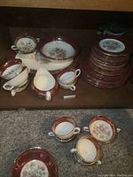 Overview of the dinnerware set showing cream soup cups, two-handled cups, saucers, and stacked plates with maroon and gold detailing and floral centers.