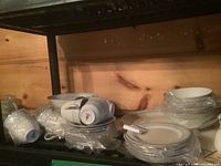 Stack of various dinnerware pieces wrapped mostly in plastic, placed on wooden shelf.