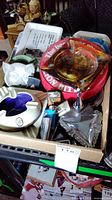 Photo of assorted ashtrays and lighters in a cardboard box, showing different styles and colors of ashtrays including clear glass, metal, and ceramic variants.