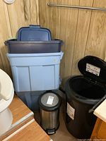Three Rubbermaid storage containers stacked, two with lids, one lid missing. Two garbage pails: one black plastic step-can with flip lid, and a small metal pedal bin with plastic lid.